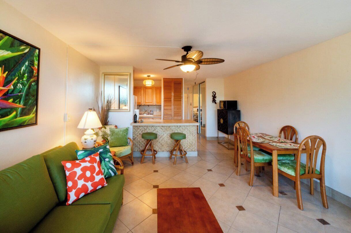 A living room with a green sofa, a dining table with chairs, a kitchen area, and a ceiling fan.