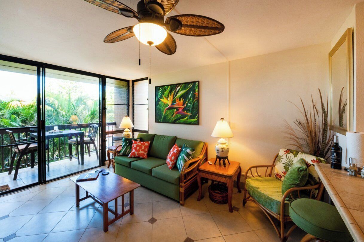 A room with a green sofa, a wooden table, a chair, a ceiling fan, and a large window with a view of greenery.