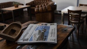 A wooden table with a newspaper, a wooden tray, and a leather couch in the background. - Hotelschiff KANAL77 - Schlafen im Hafen (Hamburg)
