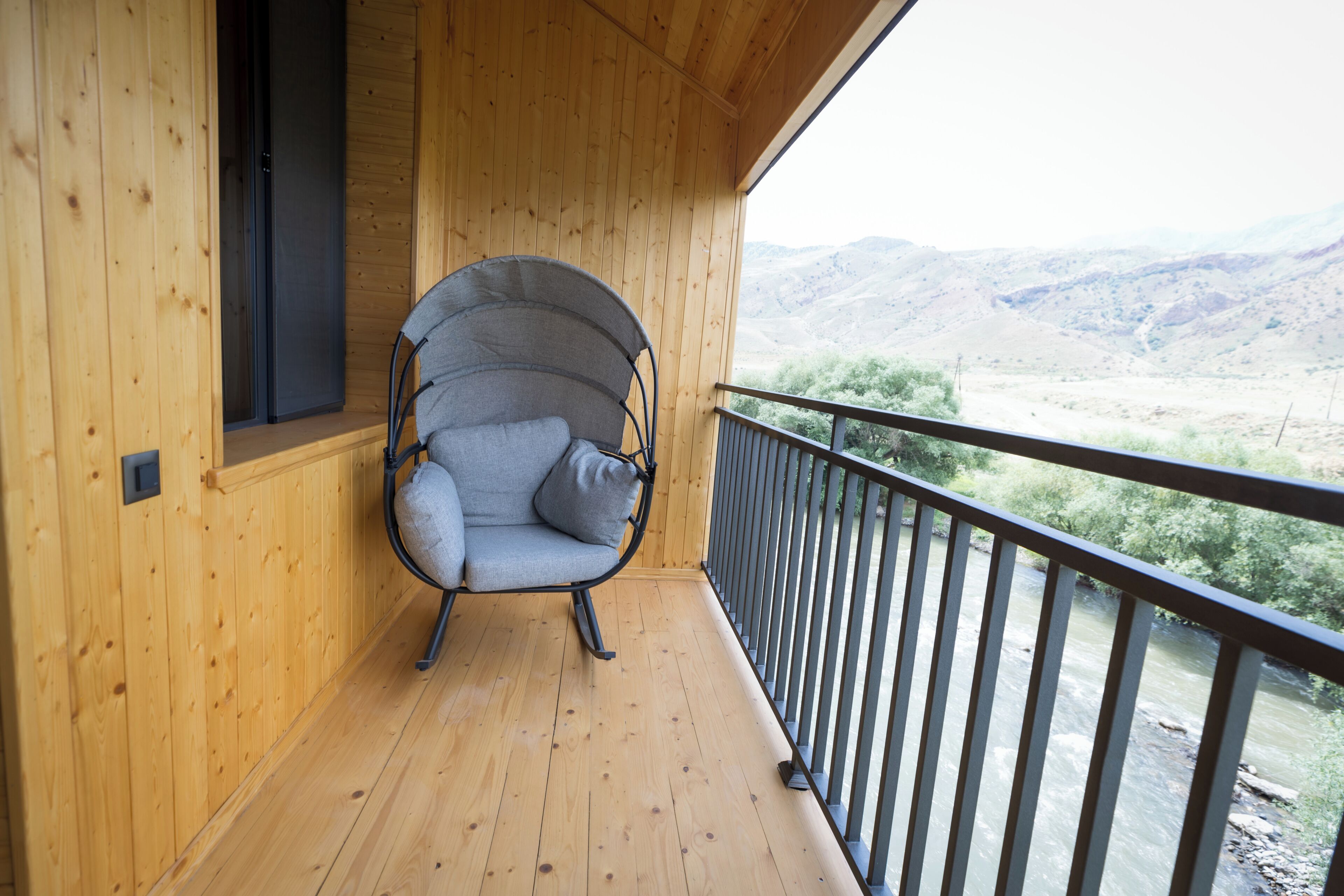 Un balcón de madera con una sola silla gris acolchada, piso de madera y vista a un río y colinas.