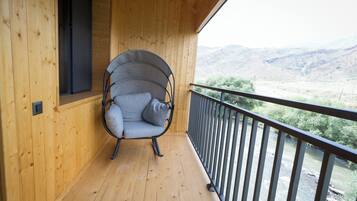 Un balcón de madera con una sola silla gris acolchada, piso de madera y vista a un río y colinas.