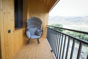 A wooden balcony with a single grey cushioned chair, a wooden floor, and a view of a river and hills. - Lchak Hotel (Yeghegnadzor)