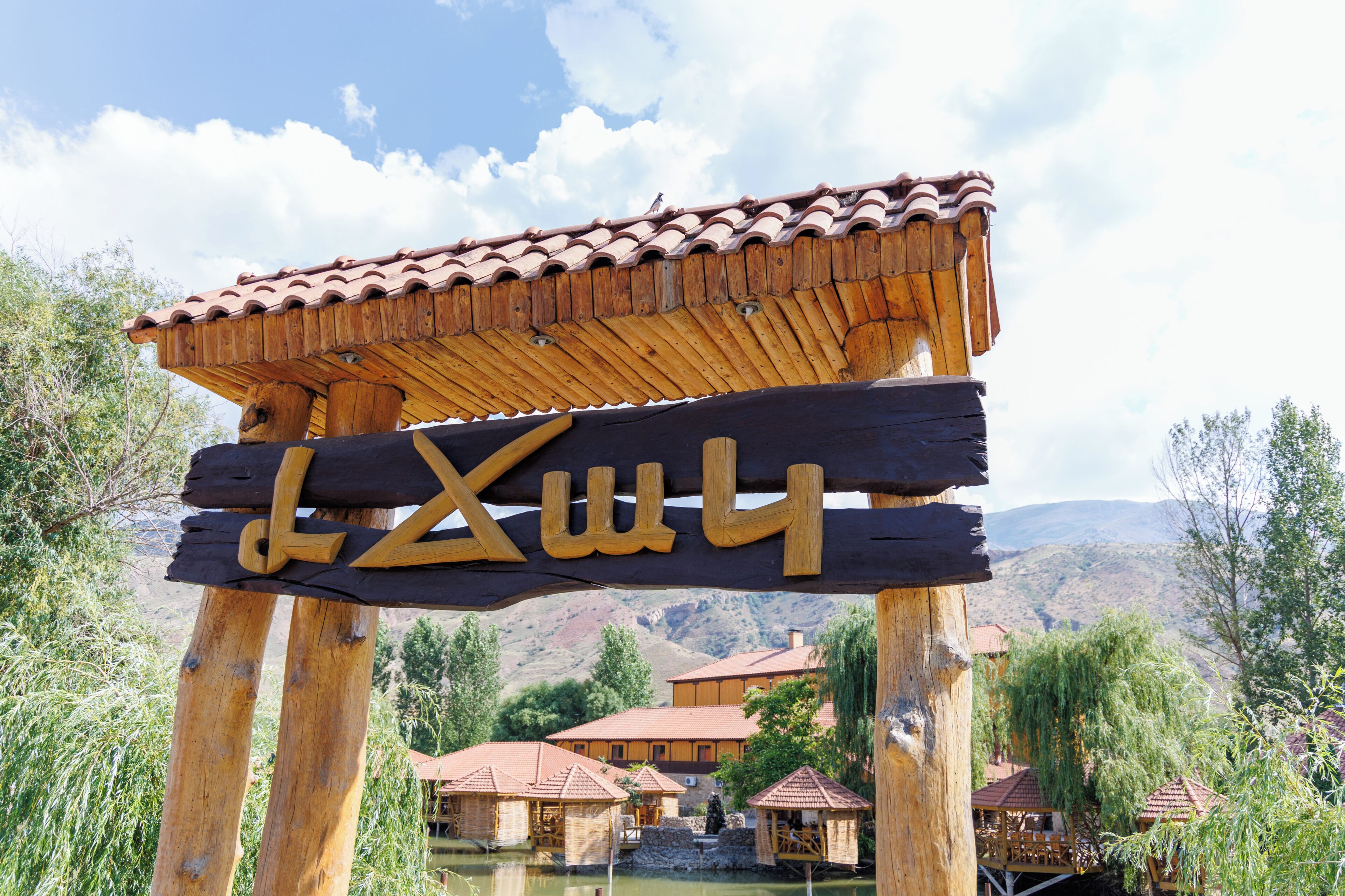A wooden sign with a tiled roof, wooden pillars, and a view of traditional buildings and a body of water in the background.