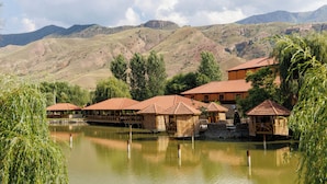 Una serie di edifici tradizionali in legno con tetti in tegole, situati in prossimità di un'acqua calma, circondati da una vegetazione rigogliosa e dolci colline sullo sfondo.