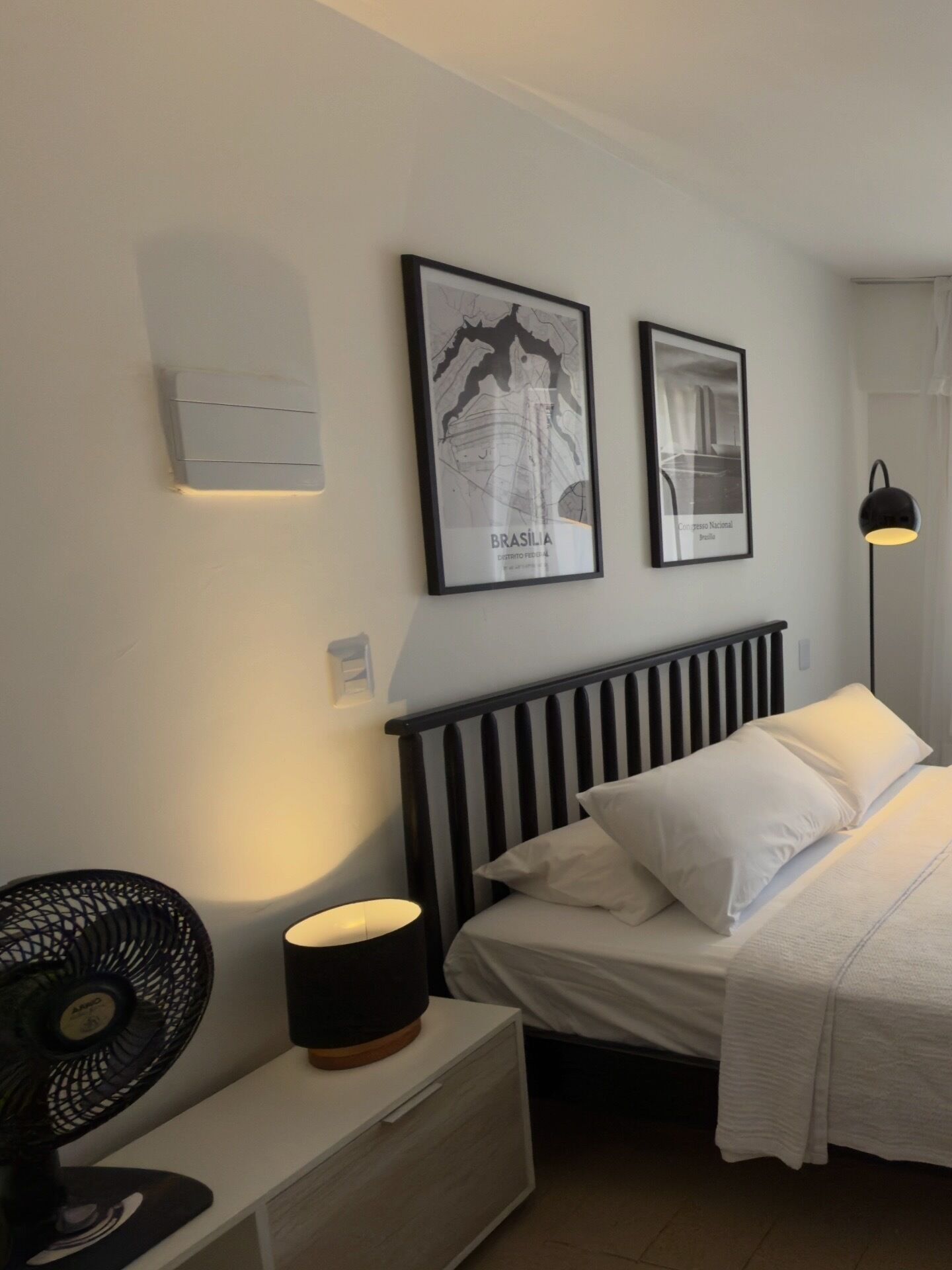 A hotel room with a bed, a nightstand, a lamp, a fan, and two framed pictures on the wall.