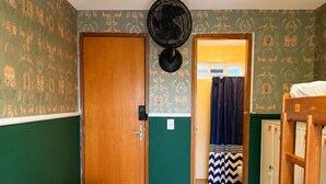 Una habitación con puerta de madera, un ventilador, un banco, una cama y un baño con cortina de ducha.