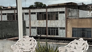 Dos sillas blancas decoradas en un patio con baldosas, con una planta en maceta entre ellas, con vista a un edificio con columna blanca y techo de metal corrugado.