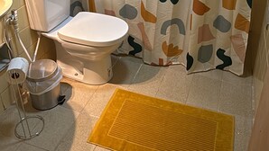 Un baño con inodoro, cortina de ducha con diseños abstractos, una alfombra amarilla y suelo de baldosas.