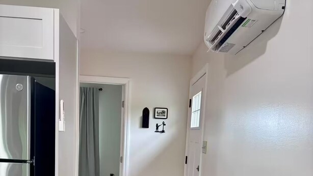 Een smalle gang met een koelkast links, een airconditioning rechts, en een deur aan het einde die leidt naar een andere kamer.