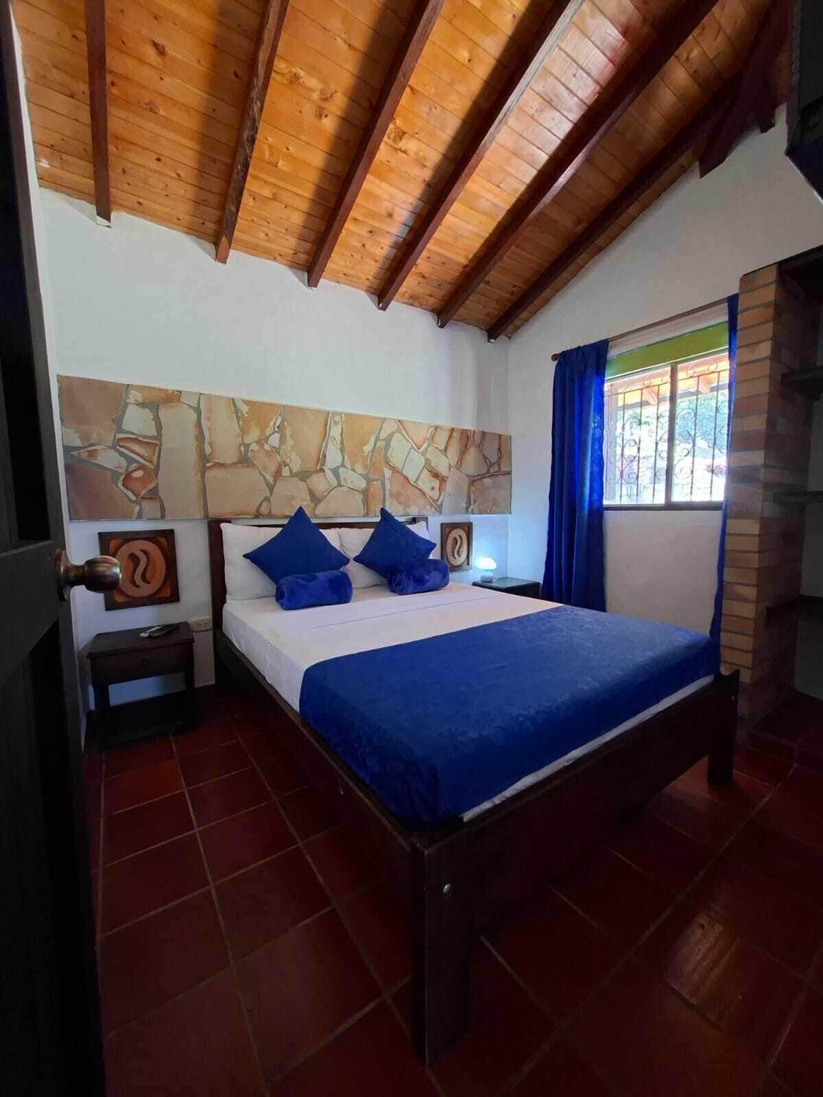 Une chambre avec un plafond en bois, une œuvre d’art murale en pierre, un lit avec une literie bleue, une table de chevet et une fenêtre avec des rideaux.