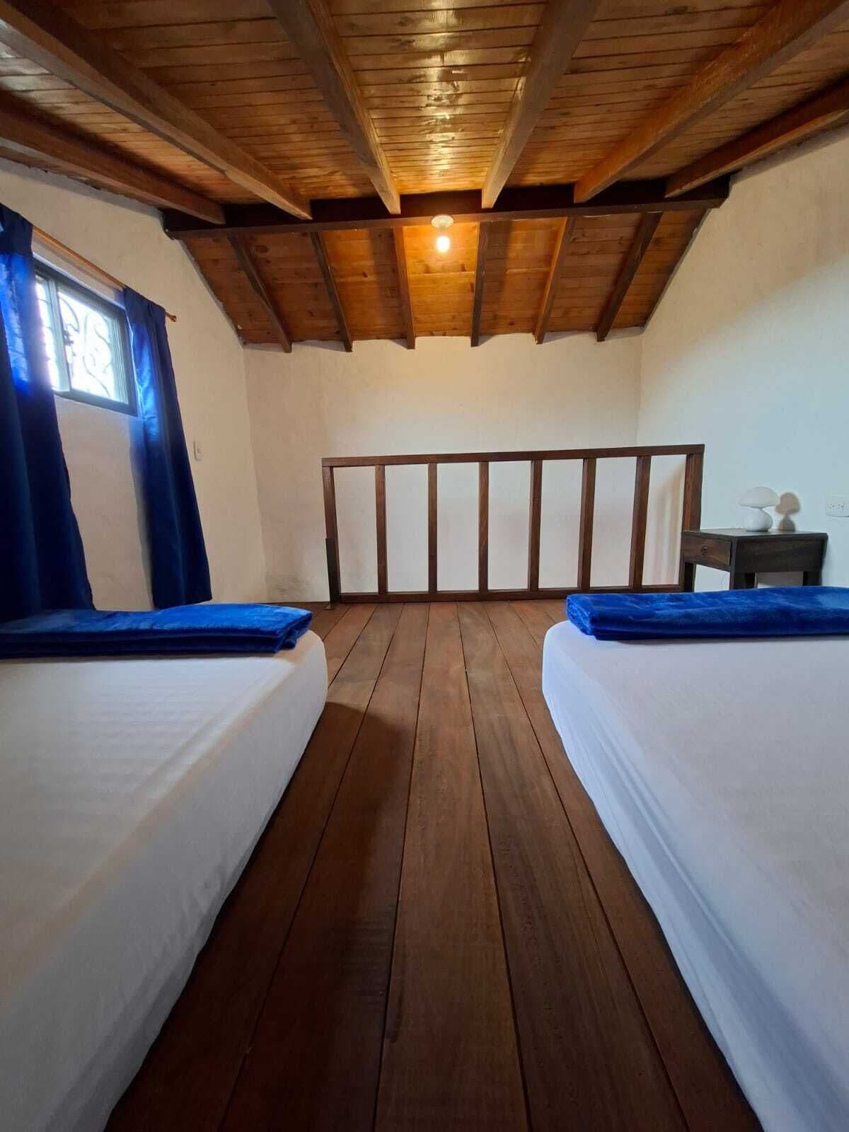 A room with two beds, wooden flooring, a sloped ceiling, and a window with curtains.