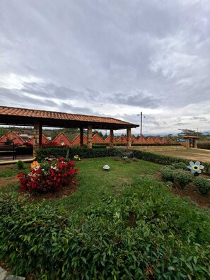 Un espacio exterior cubierto con techo de baldosas, rodeado de setos ordenados y diversas plantas florecientes.