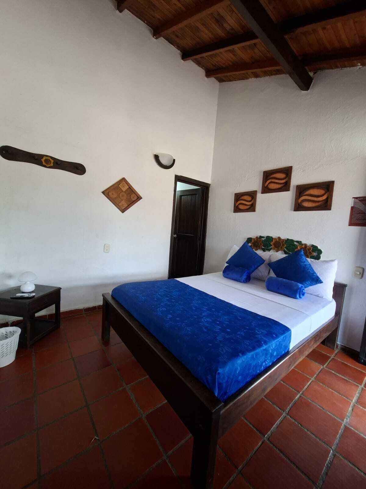 Een enkel bed met een blauwe spreidek en kussens, een houten hoofdbord, een klein nachtkastje met een lamp en een wandplank met decoratieve objecten.