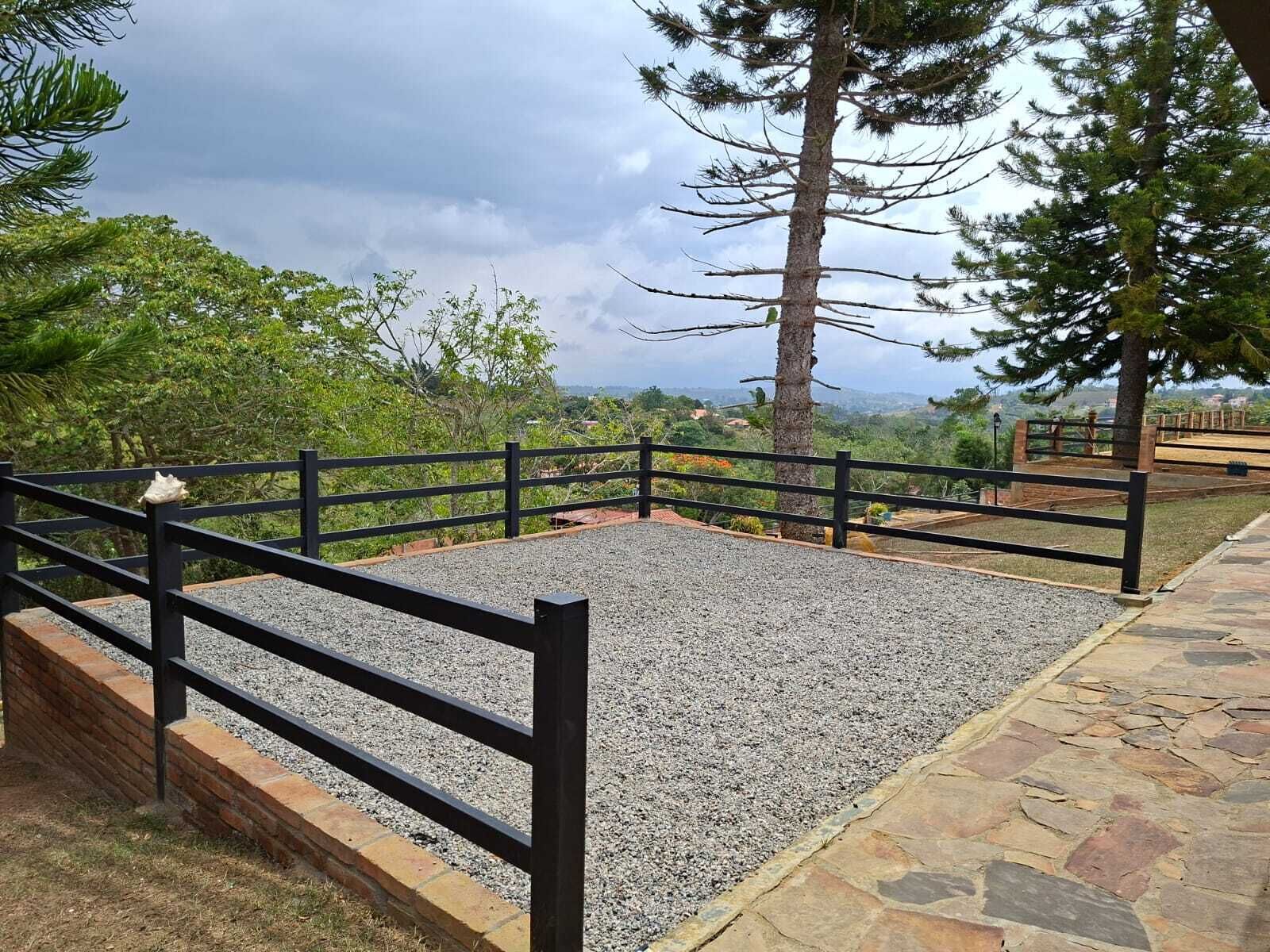 Un área pavimentada con una cerca de metal negro, rodeada de árboles y una vista panorámica del paisaje distante.