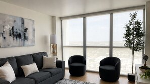 A modern living room with a grey sofa, two black armchairs, a patterned rug, a large window with a city view, and a tall plant. - Lovely 1-bedroom apartment with AC in Fort Myers (Fort Myers)