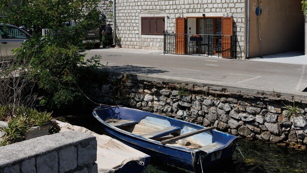 Un bâtiment en pierre surmonté d’un toit de tuiles rouges, un petit bateau amarré à un quai en pierre, et un paysage montagneux en arrière-plan.