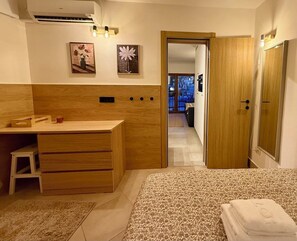 Ein Schlafzimmer mit einem hölzernen Schminktisch, einem Bett mit floralem Überwurf, einem kleinen Hocker und einer Tür zu einem anderen Raum.