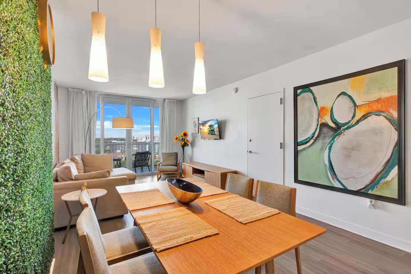 A modern hotel room with a dining area, a sofa set, a large abstract painting, and a view of the city through the window.