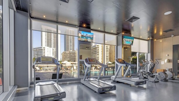 A gym with treadmills and exercise equipment, large windows, and a view of city buildings. - Sky Views 2BR  Intracoastal (Hallandale Beach)