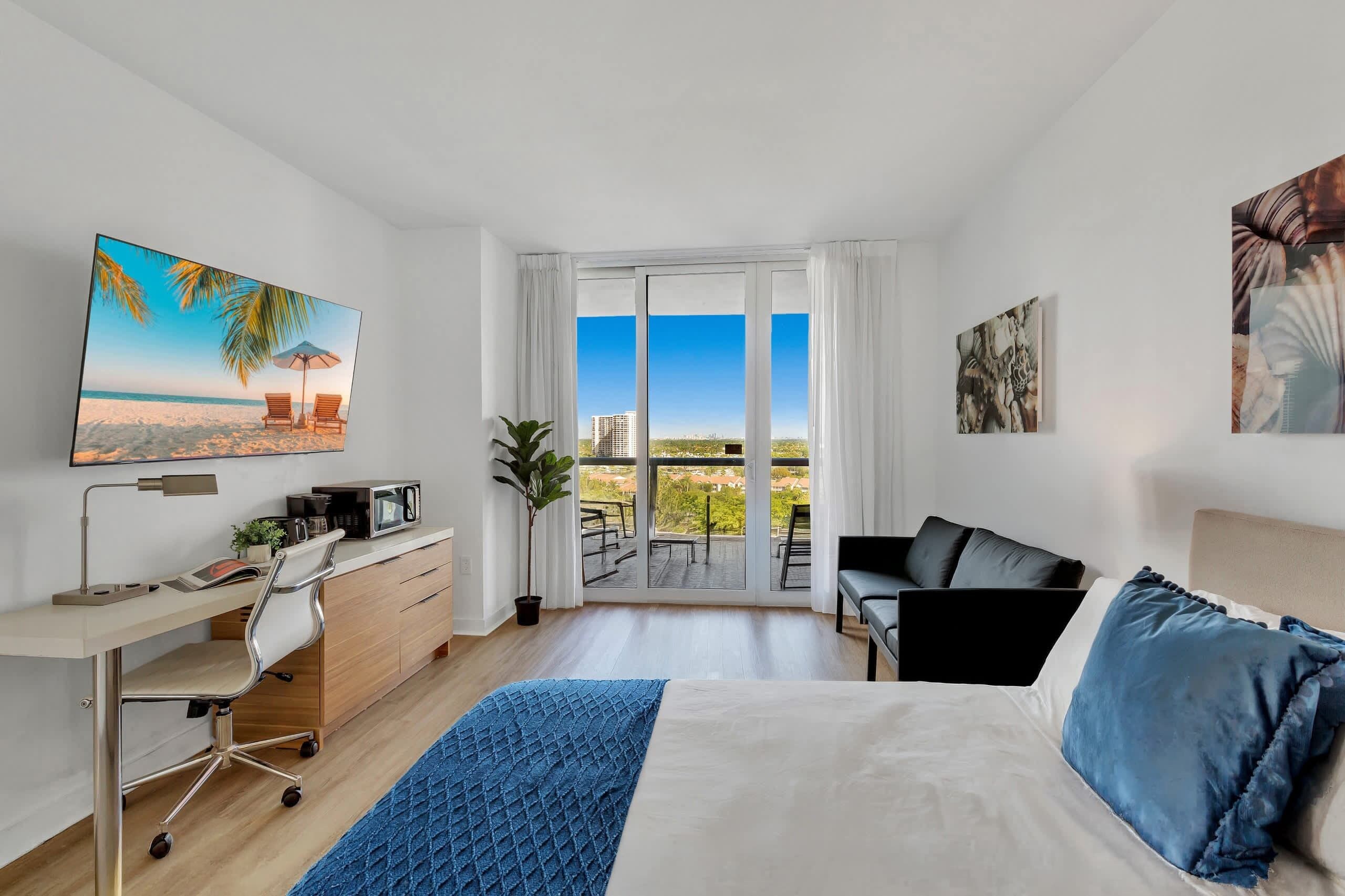 Quarto de hotel com cama, escrivaninha, cadeira, laptop, televisão e varanda com vista.