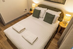 A neatly made bed with white linens and two folded towels, a bedside lamp, and a small potted plant on the nightstand. - Cosy 1BR Flat Near York Minster, Bar Walls & Station (York)