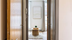 Ein Flur mit einer Holztür, einem Telefon, einem gerahmten abstrakten Kunstwerk, einem kleinen Tisch mit einer Pflanze und einem Fenster mit Vorhängen.