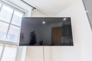 Un téléviseur à écran plat fixé au mur dans une pièce dotée d’une fenêtre, d’une plante et d’un coin cuisine visible dans le reflet.