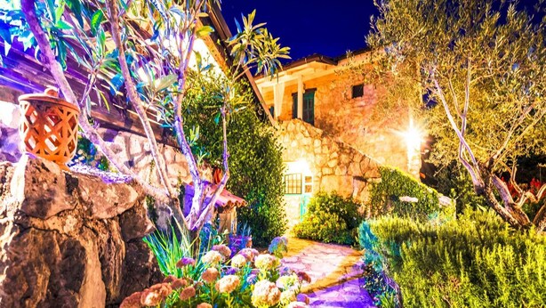 Um edifício de pedra com um caminho de jardim, rodeado de vegetação e iluminado por luzes artificiais.