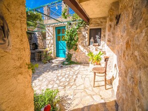 Une cour pavée de pierres, avec une porte bleue, un pot de fleurs et une chaise en bois.