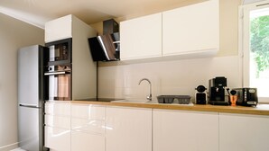 Une cuisine moderne avec des meubles blancs, un réfrigérateur en acier inoxydable, un four encastré, une machine à café et une fenêtre laissant entrer la lumière naturelle.