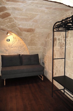 A stone-walled room with a dark wooden floor, a grey sofa with cushions, and a black metal shelf unit.