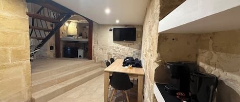 A stone-walled room with a wooden table and chairs, a television mounted on the wall, and a staircase leading to an upper level.