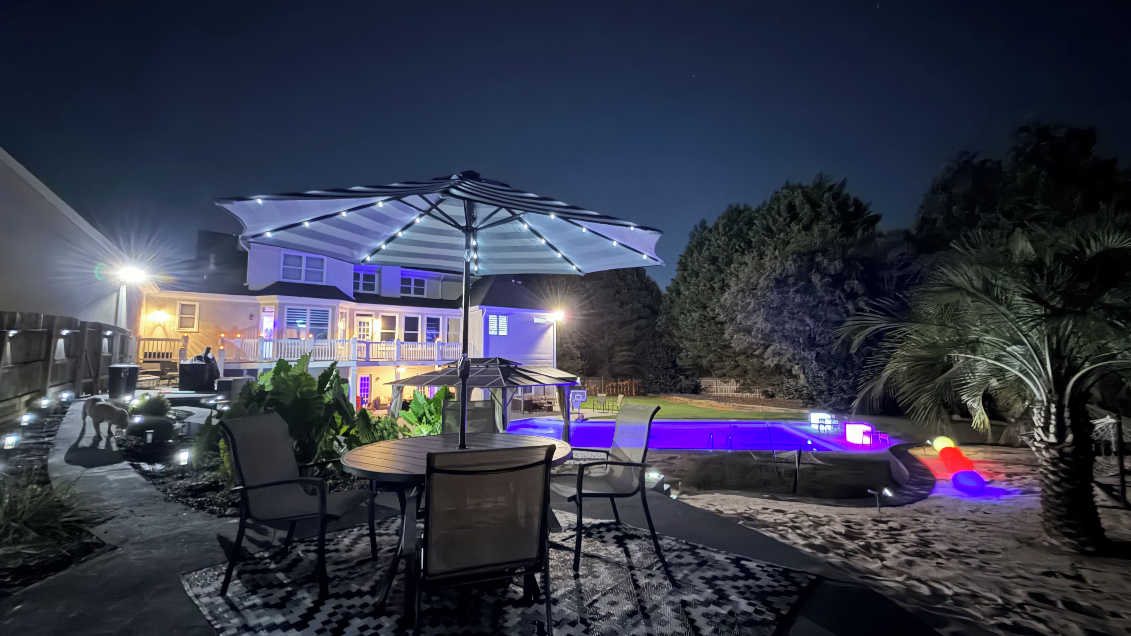 A patio area with a pool, outdoor furniture, and a house with illuminated windows.