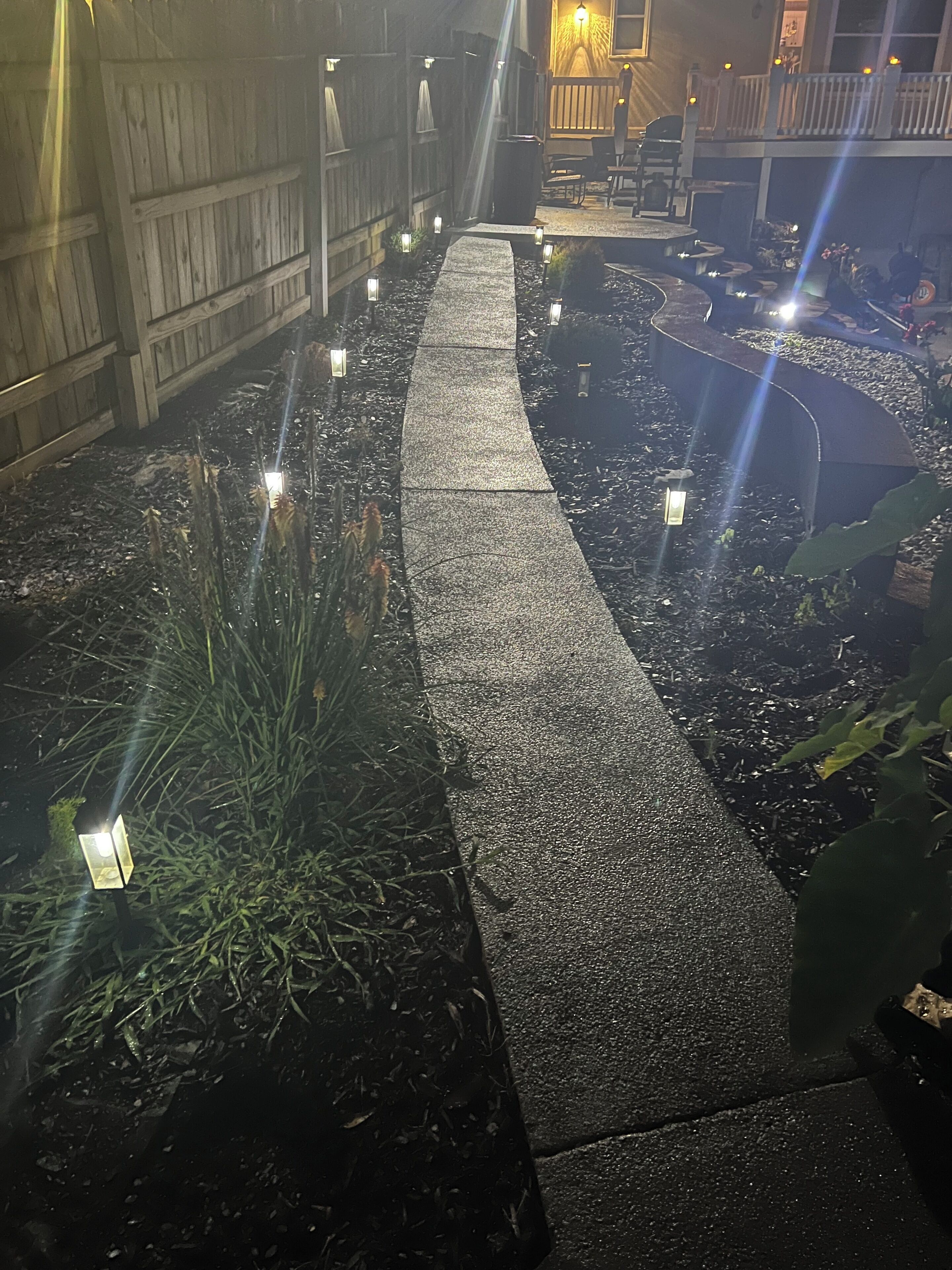 A garden pathway with stepping stones and small lights, surrounded by plants and a wooden fence.