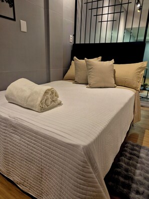 A neatly made bed with a folded white towel, a headboard with multiple pillows, and a modern design. - Cozy studio in downtown Florianópolis. Close to everything the island has to offer. (Florianópolis)