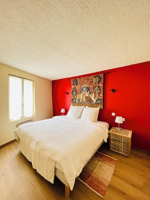 A bedroom with a large bed, a red accent wall, a window, and a nightstand with a lamp. - A 3-story, 110-square-meter house from the 17th century at the foot of the Cité de Carcassonne (Carcassonne)
