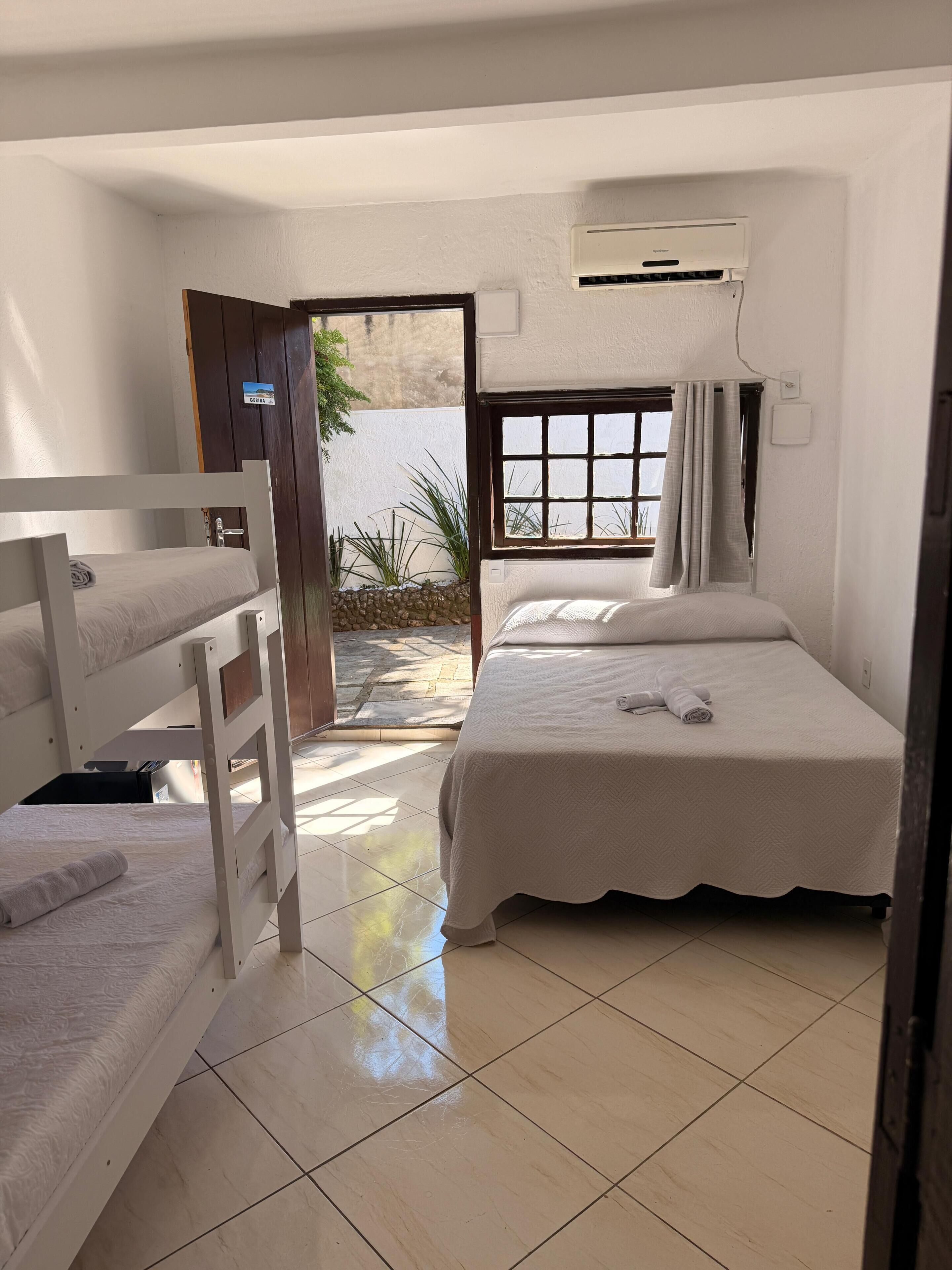 Quarto com beliches, janela com cortinas, piso em azulejos e porta que dá para o exterior.