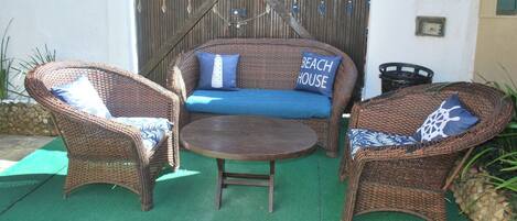 Un espace patio aménagé avec des meubles en osier, comprenant deux chaises et un canapé agrémenté d’un coussin portant l’inscription « MAISON DE PLAGE ».