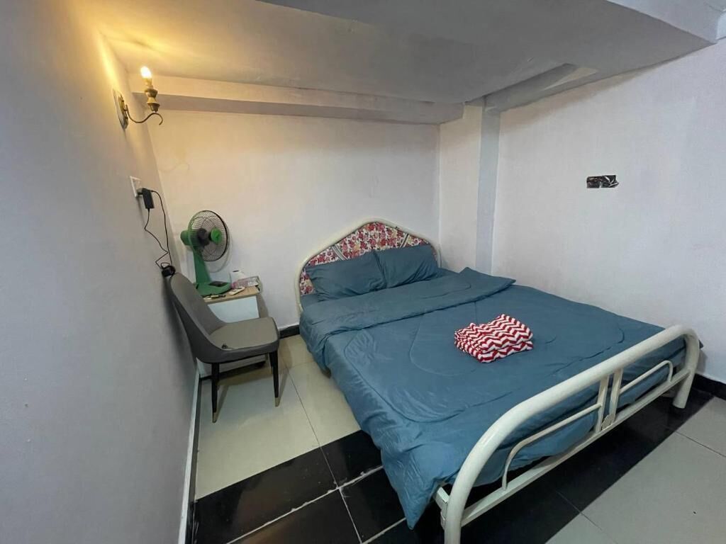 Una cama individual con estructura metálica, una mesita de noche con lámpara y un ventilador de pared.