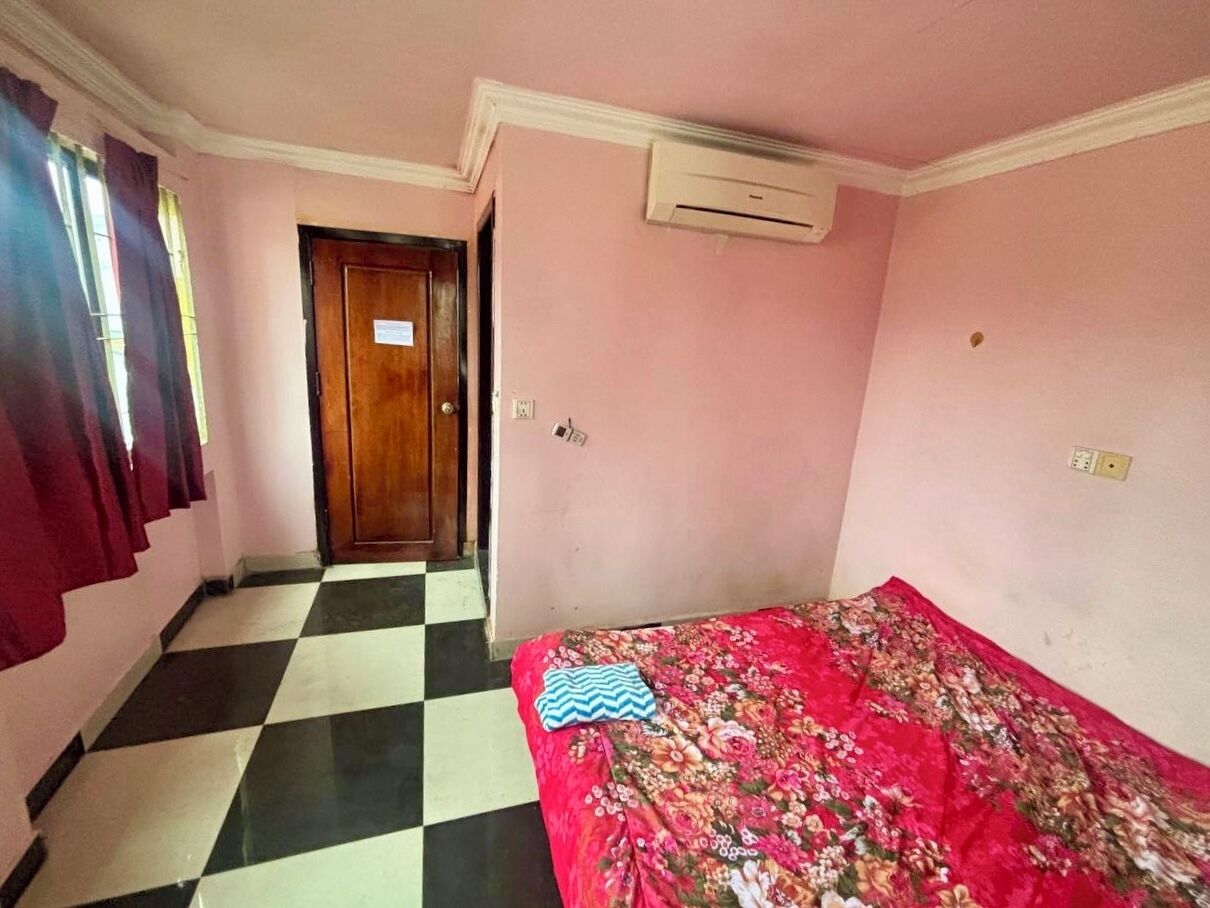 A room with a checkered floor, a bed with a floral bedspread, a wooden door, and an air conditioning unit.