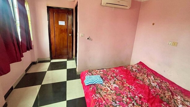 A room with a checkered floor, a bed with a floral bedspread, a wooden door, and an air conditioning unit. - Arecrata (Phnom Penh)