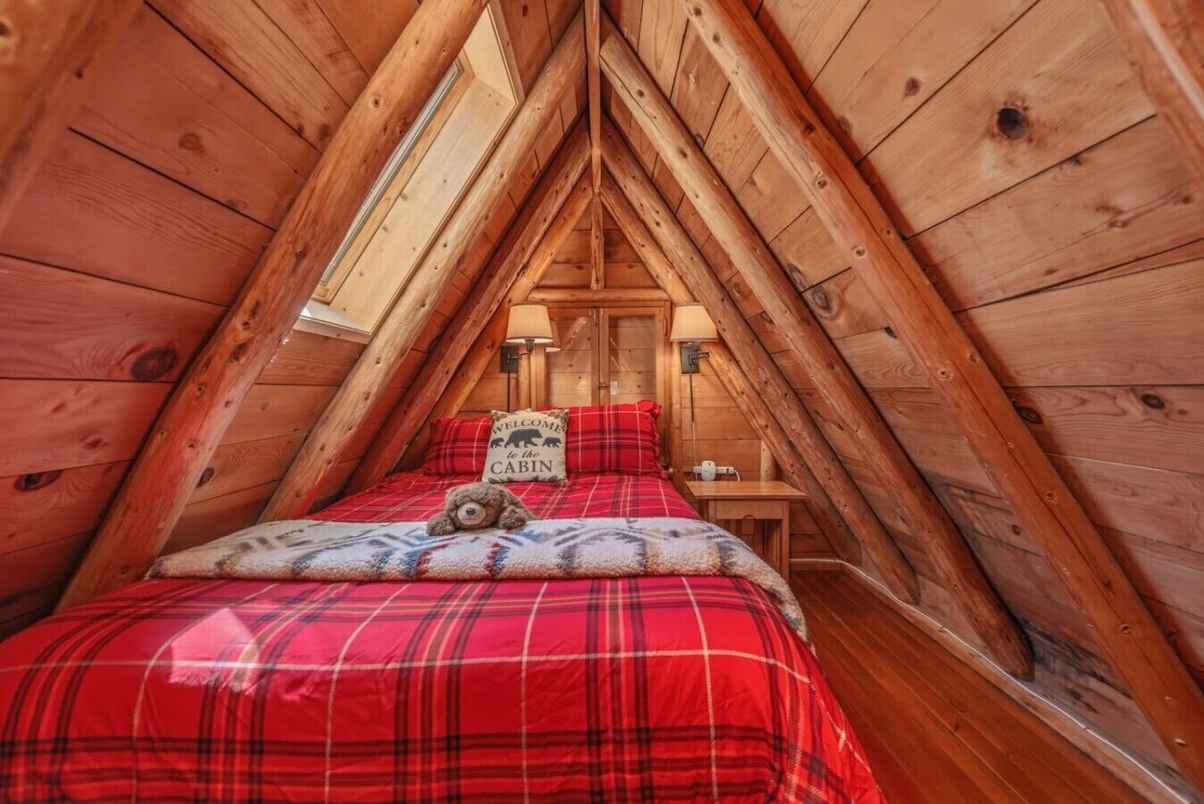 A cozy attic bedroom with a sloped ceiling, a bed with a plaid bedspread, a welcome pillow, and a teddy bear.