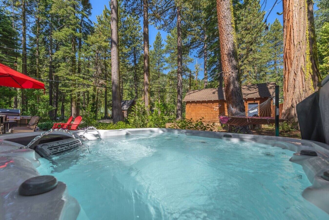 A hot tub surrounded by tall trees, a wooden cabin, and red chairs.