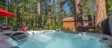 A hot tub surrounded by tall trees, a wooden cabin, and red chairs.