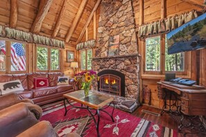 Interior de una cabaña de troncos con chimenea de piedra, muebles de cuero y un escritorio con una computadora.