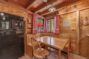 A cabin interior with a wooden table, chairs, a refrigerator, and a window with red curtains. - Dancing Cubs Cabin!  Hot Tub, Walk to Beach! (Homewood)