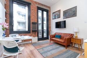 A modern living room with a brick wall, a leather sofa, a dining table with chairs, a TV mounted on the wall, and a large window. - Chic Heritage Studio in Downtown Victoria (Victoria)
