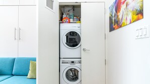 Uma lavanderia compacta com máquina de lavar e secadora frontais, um sofá azul e um quadro abstrato colorido na parede.