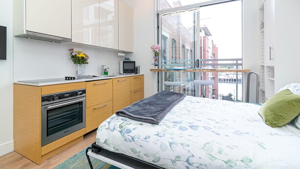 Quarto moderno com cama, uma kitchenette pequena e uma janela grande com vista para edifícios.