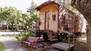 A vintage train car with a bench and a small table in front, set in a park-like area with trees and a paved path. - London Calling Railroad Caboose Cabin (Upper Lake)